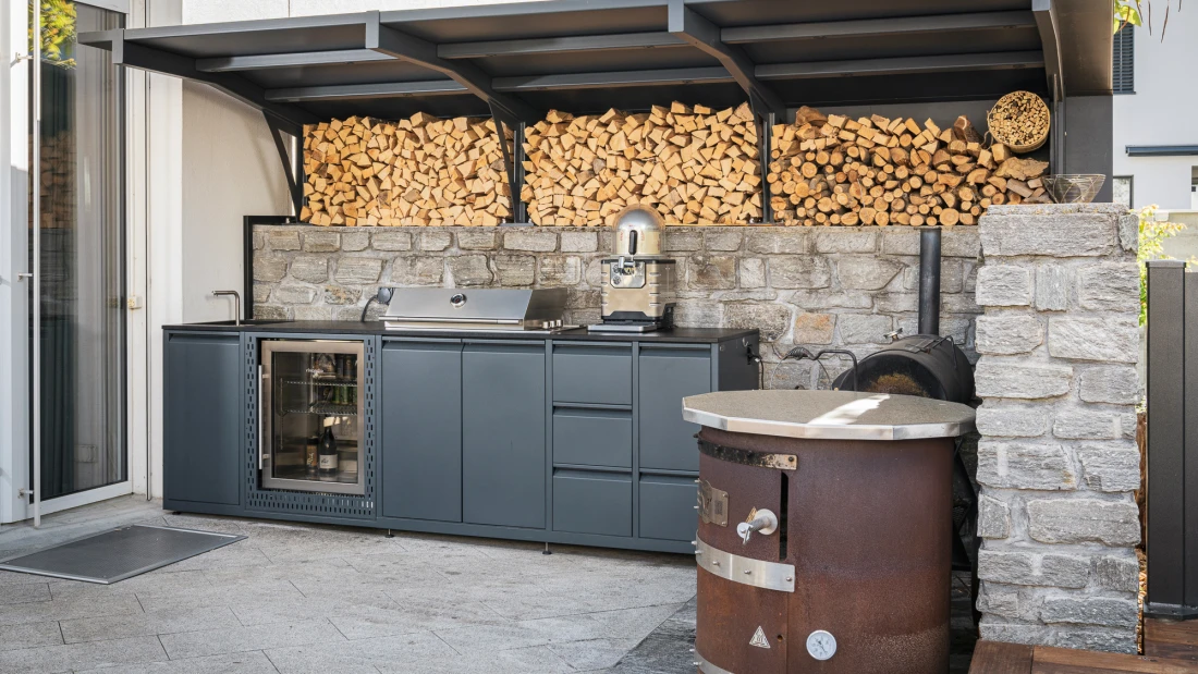 Moderne Outdoorküche mit Kühlschrank und Grill vor einer Natursteinmauer in Domat/Ems.