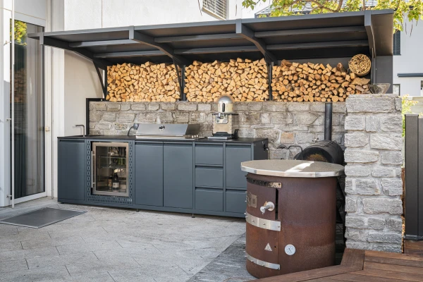 Moderne Outdoorküche mit Kühlschrank und Grill vor einer Natursteinmauer in Domat/Ems.